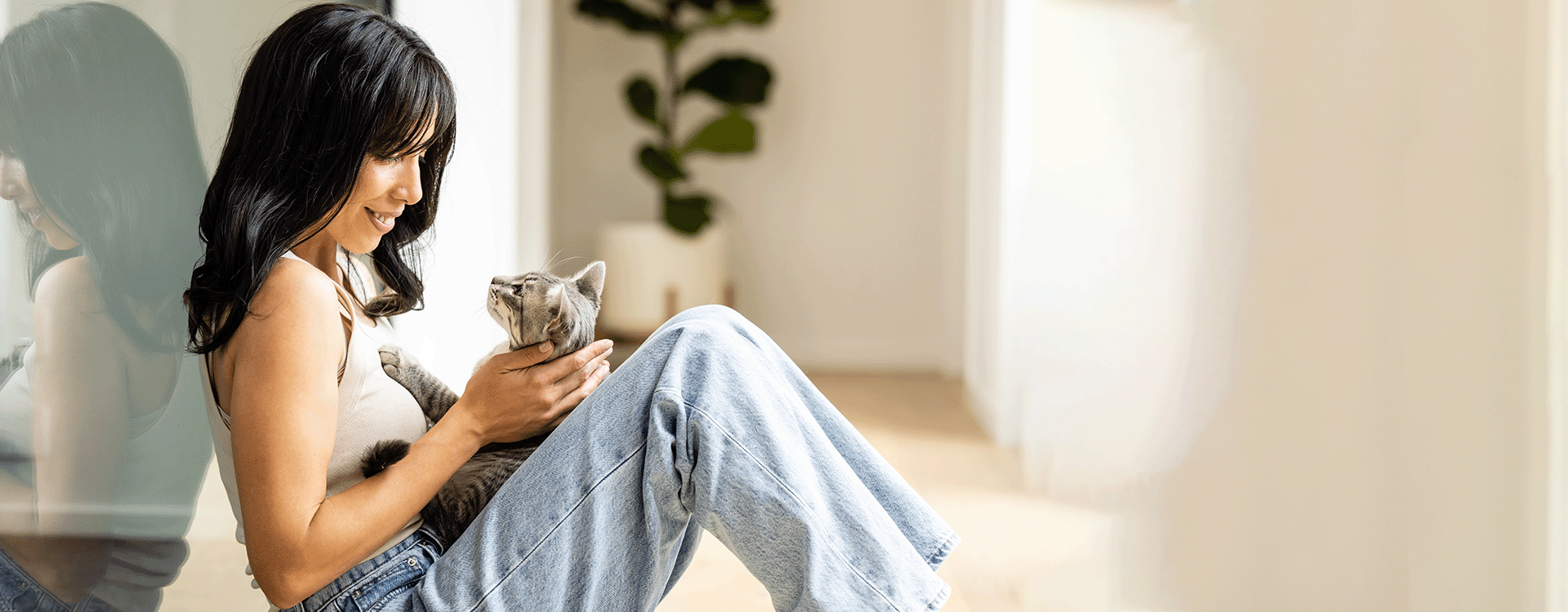 Landscape image of woman sitting on the floor with her cat on her lap. She is leaned up against a window. 