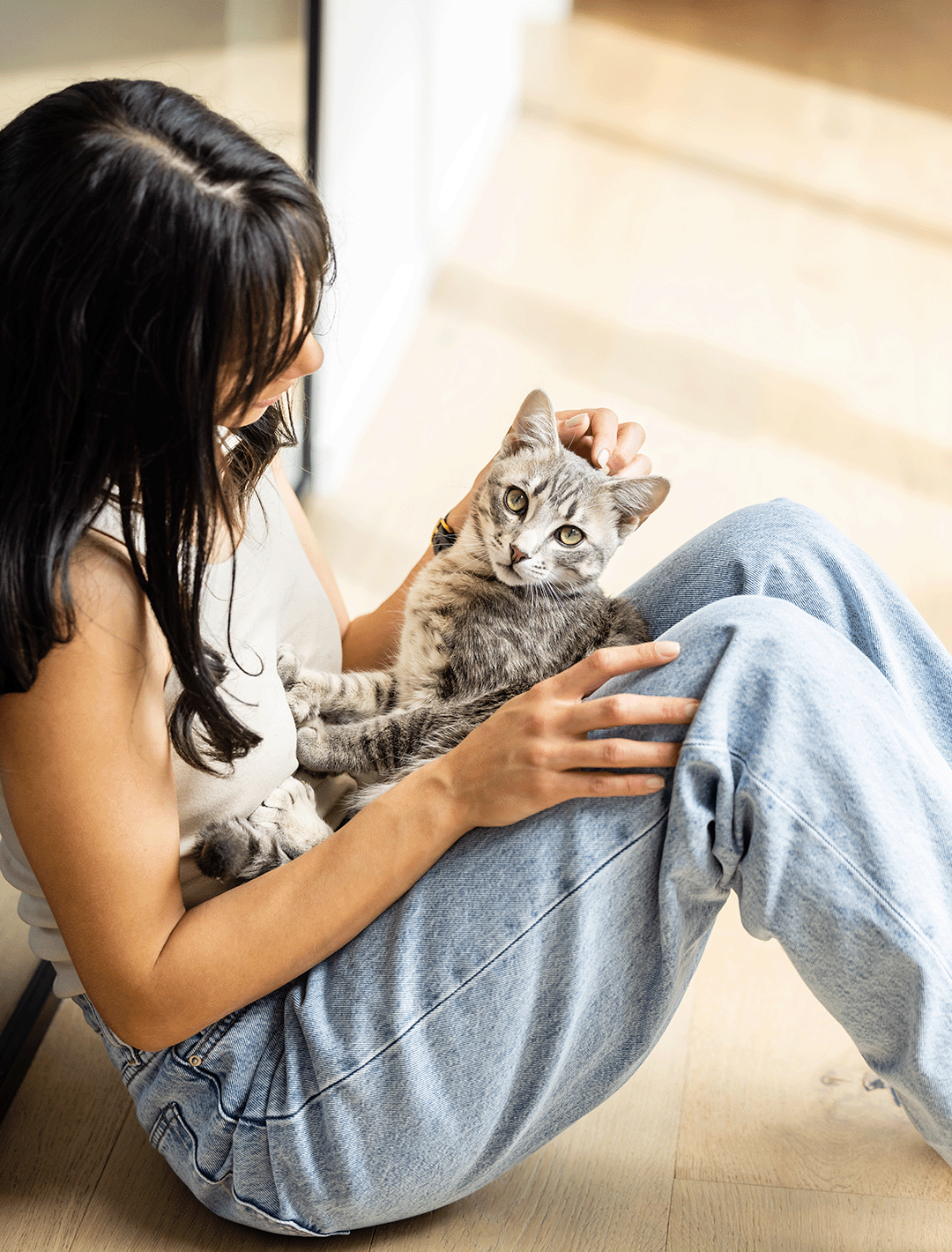 Portrait image of woman sitting on the floor with her cat on her lap. She is leaned up against a window. 